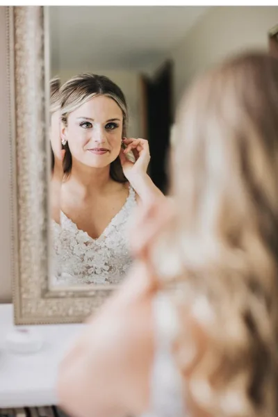 A bride in a white lace wedding dress looking into a mirror while adjusting her hair and earrings.
