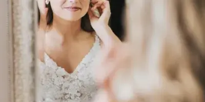 A bride in a white lace wedding dress looking into a mirror while adjusting her hair and earrings.