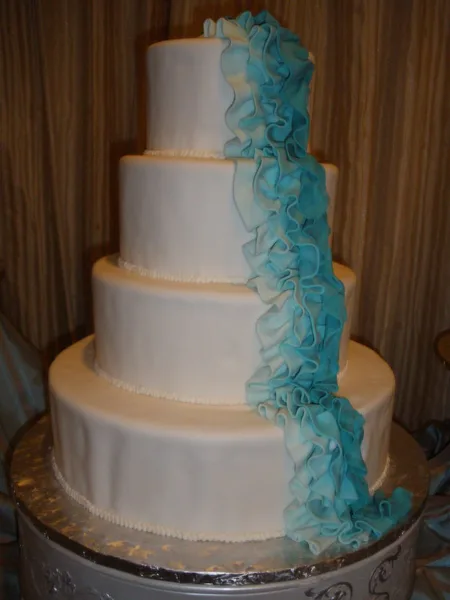 A four-tier white wedding cake decorated with cascading blue fondant ruffles.