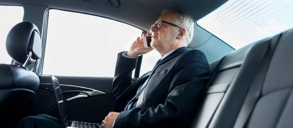 A man in a suit using a phone and laptop in the back seat of a luxury car.