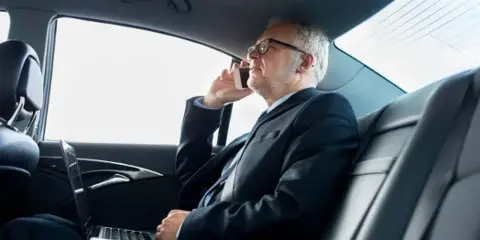 A man in a suit using a phone and laptop in the back seat of a luxury car.