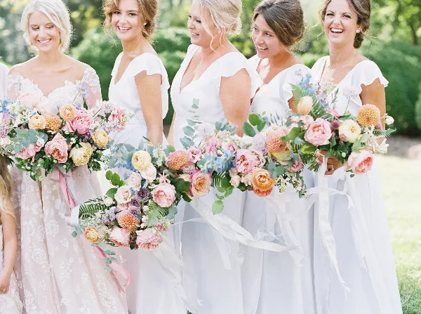 Bridesmaids in white dresses holding large, colorful wedding bouquets with pink and peach flowers and greenery.