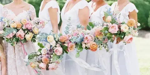 Bridesmaids in white dresses holding large, colorful wedding bouquets with pink and peach flowers and greenery.