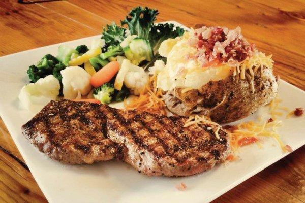 A plated meal consisting of a grilled steak, a loaded baked potato with toppings, and a side of mixed vegetables on a white square plate.