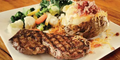 A plated meal consisting of a grilled steak, a loaded baked potato with toppings, and a side of mixed vegetables on a white square plate.