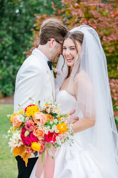 A bride and groom smiling and embracing outdoors, with the bride holding a vibrant bouquet.