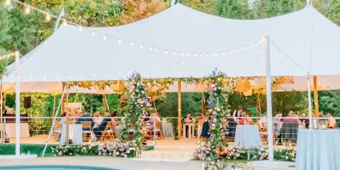 A white event tent set up outdoors for a wedding reception with floral decorations and string lights.