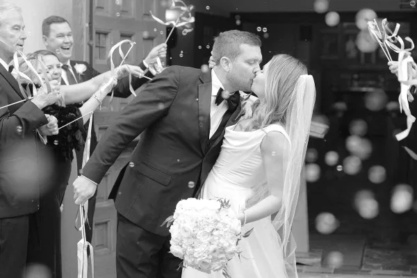 Black and white photograph of a bride and groom kissing as guests hold ribbon wands and bubbles float in the air.