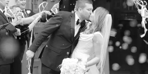 Black and white photograph of a bride and groom kissing as guests hold ribbon wands and bubbles float in the air.