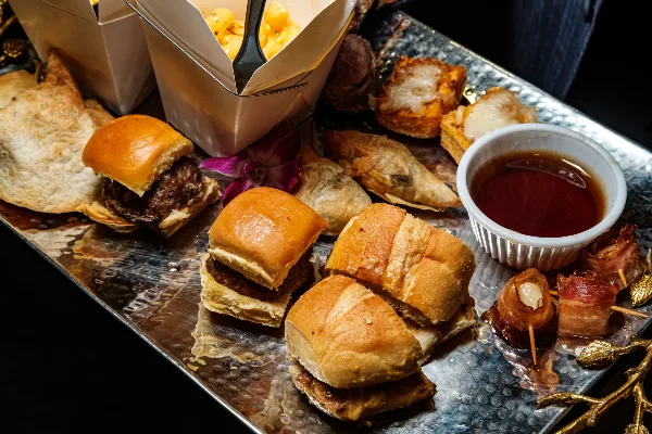 A silver tray filled with assorted catering appetizers including sliders, bacon-wrapped items, and a dipping sauce.