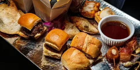 A silver tray filled with assorted catering appetizers including sliders, bacon-wrapped items, and a dipping sauce.