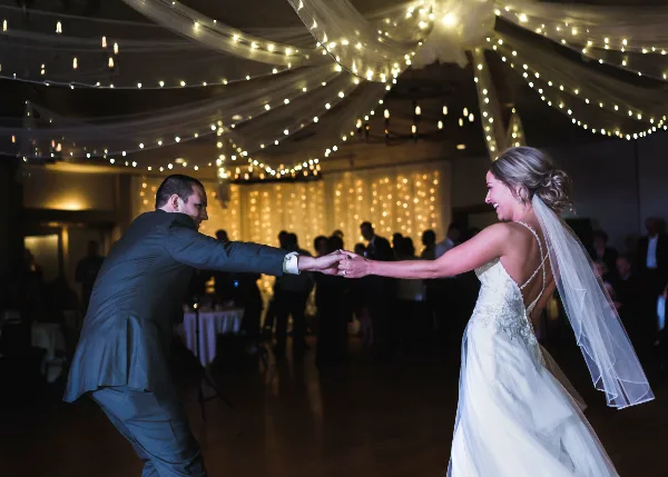 A bride and groom dancing at a wedding reception under string lights and draped ceiling decor.