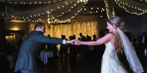 A bride and groom dancing at a wedding reception under string lights and draped ceiling decor.