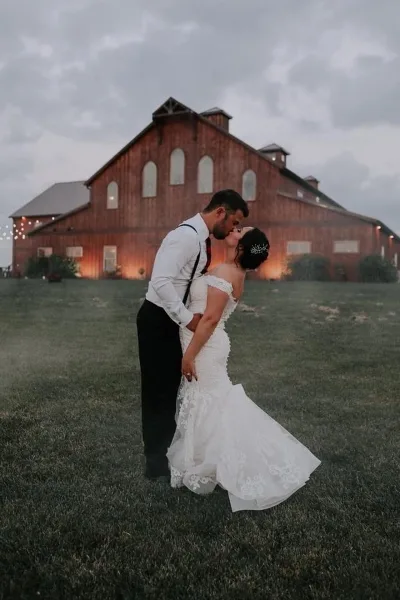 A bride and groom kissing in a grassy field in front of a large red timber-frame barn.