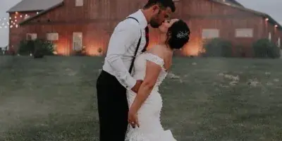 A bride and groom kissing in a grassy field in front of a large red timber-frame barn.
