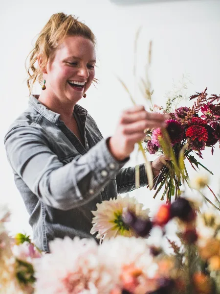 A smiling woman in a denim shirt arranging a colorful bouquet of flowers in a studio setting.