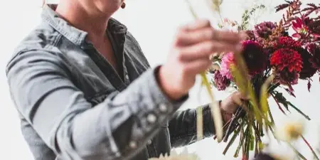 A smiling woman in a denim shirt arranging a colorful bouquet of flowers in a studio setting.