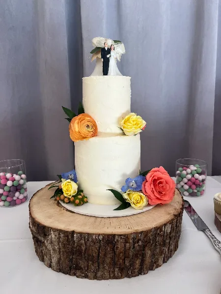 A two-tier white wedding cake decorated with orange, yellow, and pink flowers, topped with a bride and groom figurine, sitting on a rustic wooden slab.