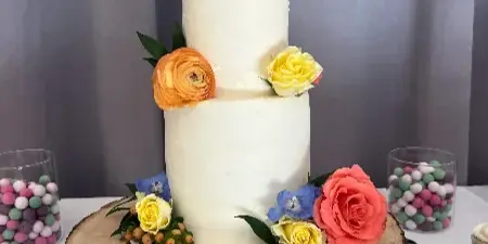 A two-tier white wedding cake decorated with orange, yellow, and pink flowers, topped with a bride and groom figurine, sitting on a rustic wooden slab.