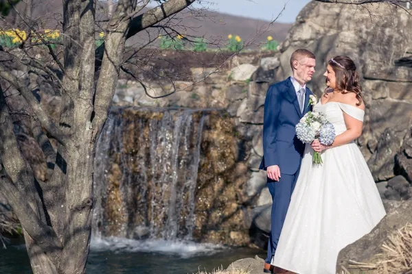 A bride and groom standing together outdoors near a waterfall, with the bride holding a bouquet of white flowers.