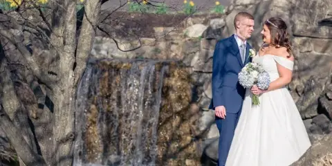 A bride and groom standing together outdoors near a waterfall, with the bride holding a bouquet of white flowers.