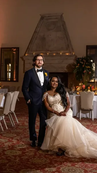 A bride and groom posing together in a formal event hall with floral arrangements and candlelight.