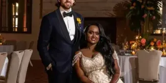 A bride and groom posing together in a formal event hall with floral arrangements and candlelight.