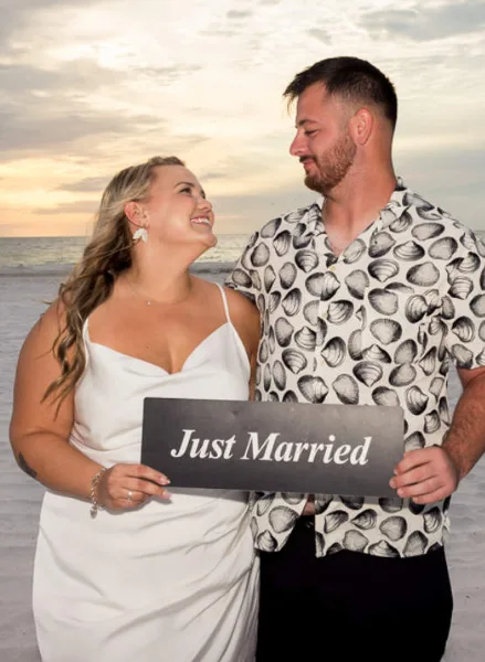 A bride and groom standing on a beach at sunset holding a 'Just Married' sign.