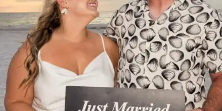 A bride and groom standing on a beach at sunset holding a 'Just Married' sign.