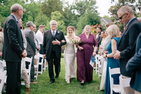 A bride in a white gown walks down the aisle between rows of seated guests during an outdoor wedding ceremony.