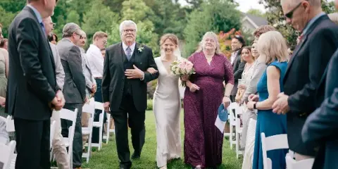 A bride in a white gown walks down the aisle between rows of seated guests during an outdoor wedding ceremony.