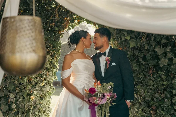 Bride and groom in wedding attire standing together at a leafy ceremony arch with a floral bouquet.