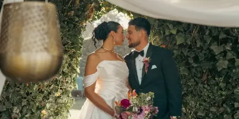 Bride and groom in wedding attire standing together at a leafy ceremony arch with a floral bouquet.