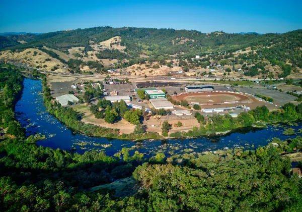 Aerial view of the Douglas County Fairgrounds Complex showing the oval racetrack, buildings, and parking lot surrounded by a river and forested hills.