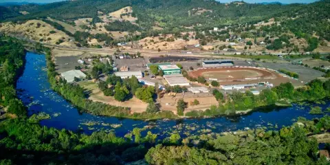 Aerial view of the Douglas County Fairgrounds Complex showing the oval racetrack, buildings, and parking lot surrounded by a river and forested hills.