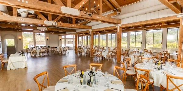 Interior of a rustic event venue with wooden beams, chandeliers, and round tables set for a wedding or banquet.