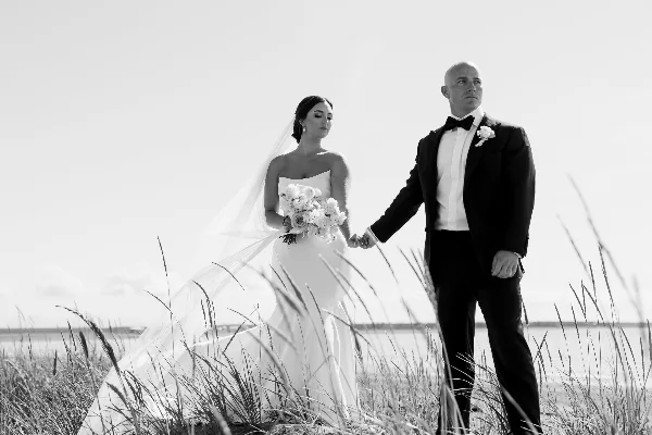 A black and white editorial wedding photograph of a bride and groom holding hands in a field of tall grass.
