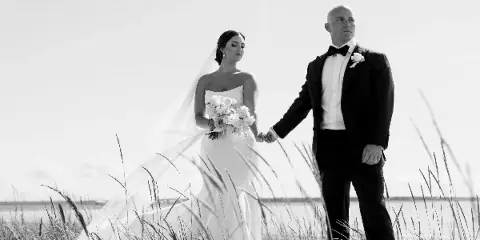 A black and white editorial wedding photograph of a bride and groom holding hands in a field of tall grass.
