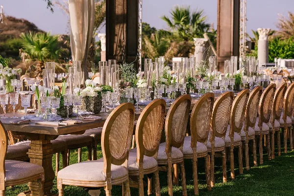 An outdoor event table setting featuring wooden chairs, glassware, and floral centerpieces.