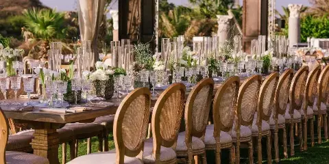 An outdoor event table setting featuring wooden chairs, glassware, and floral centerpieces.