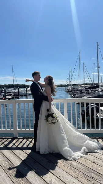 A bride and groom embracing on a wooden dock at a marina with sailboats in the background.