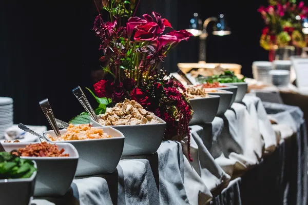 A catering buffet table with bowls of food, serving spoons, and a floral centerpiece.