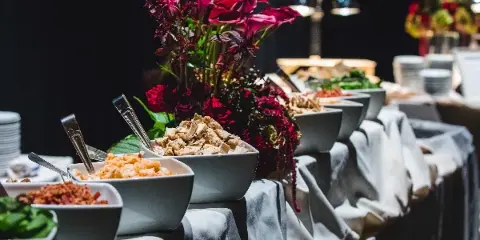 A catering buffet table with bowls of food, serving spoons, and a floral centerpiece.