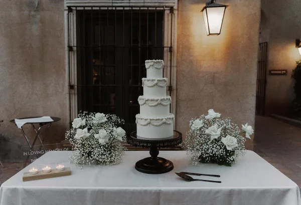 A four-tier white wedding cake with ruffled piping details displayed on a dark pedestal stand, flanked by white rose and baby's breath arrangements on a table.