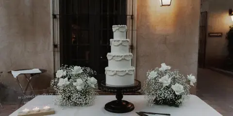 A four-tier white wedding cake with ruffled piping details displayed on a dark pedestal stand, flanked by white rose and baby's breath arrangements on a table.