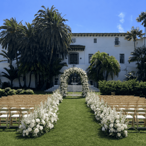 An outdoor wedding ceremony setup featuring rows of wooden folding chairs, a white floral arch, and floral aisle arrangements on a lawn.