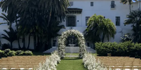 An outdoor wedding ceremony setup featuring rows of wooden folding chairs, a white floral arch, and floral aisle arrangements on a lawn.