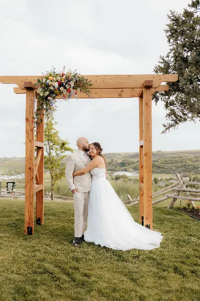 A bride and groom embracing under a wooden wedding arch with floral decor at an outdoor venue.