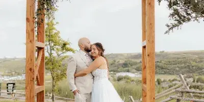 A bride and groom embracing under a wooden wedding arch with floral decor at an outdoor venue.