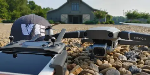 A DJI Mavic 2 drone and controller with a VE Videography cap on a gravel surface in front of a house.
